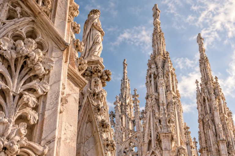Carved marble figures and Gothic pinnacles on Milan’s Duomo beneath a pale blue sky; editorial illustration.